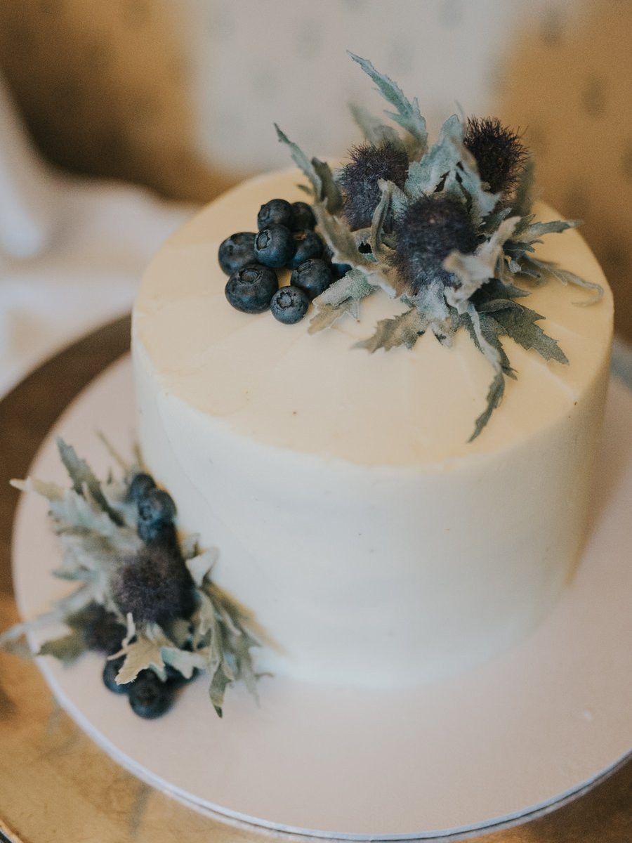 Zweistöckige Hochzeitstorte mit weißem Fondant, dekoriert mit Blaubeeren und schottischen Disteln auf silbernem Tortenteller.