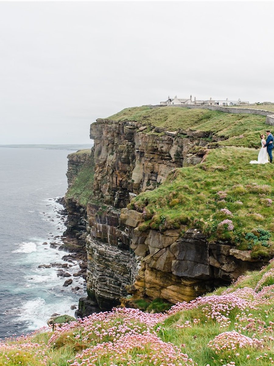 Brautpaar steht auf den Klippen von Dunnet Head an der Nordküste Schottlands, unter sich das offene Meer und wilde blühende Grasnelken.